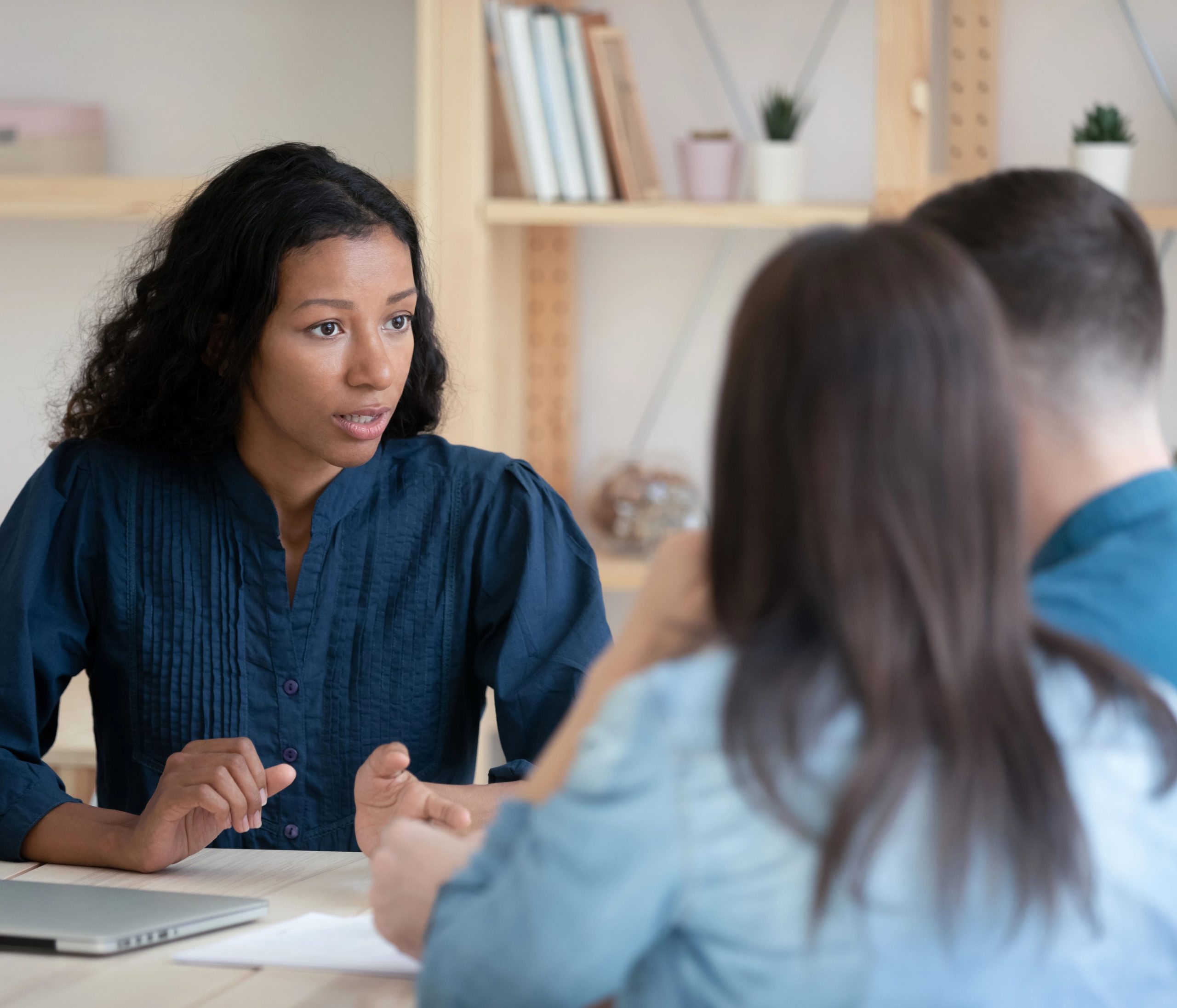 A woman with dark, curly hair wearing a blue shirt speaks to two people across a table.