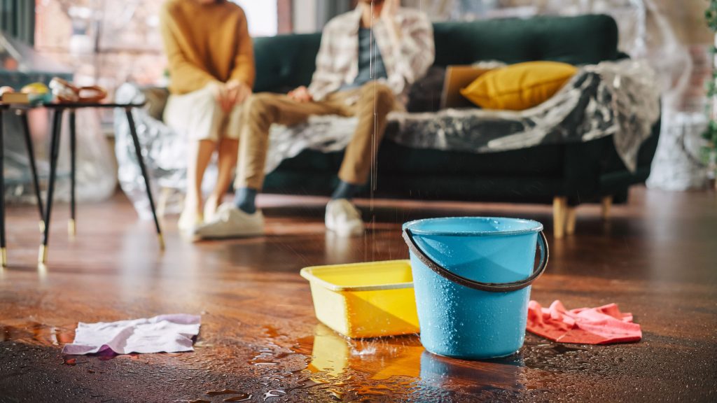 A blue bucket and a yellow basin on a wooden floor collecting water from a ceiling leak, with a soaked cloth nearby and a worried couple sitting on a couch in the background.