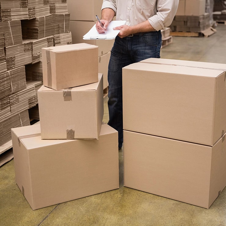 A worker in a warehouse checking inventory on a clipboard, surrounded by stacked cardboard boxes and bundles of folded cardboard.